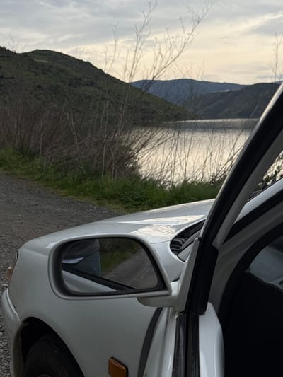 A white compact car is positioned on a gravel track, with its front passenger side facing the viewer and its passenger door ajar. The vehicle's side mirror captures a partial reflection of a person, displaying a torso clad in a grey knitted garment and light-colored trousers. The immediate foreground consists of a gravel surface bordered by green vegetation and sparse, bare tree branches. Directly behind this vegetation lies a calm body of water, possibly a river or reservoir, reflecting the diffused light from an overcast sky. The background features multiple layers of hills and mountains, densely covered in vegetation, receding into the distance under a cloudy sky. The landscape, characterized by significant water bodies and hilly terrain, is consistent with the natural environment found in and around Mogadouro, Portugal, particularly near the Douro River or its tributaries. No specific landmarks for pinpointing a precise location within Mogadouro are visible.