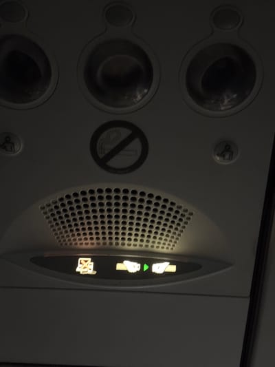 The image shows the interior of an airplane cabin, specifically the overhead panel. There are three circular air vents at the top. Below these vents, on the left, is a symbol of a person raising their hand, likely indicating a call button. In the center, there is a no-smoking symbol. On the right, there is another symbol of a person raising their hand, also likely a call button. Below the no-smoking sign is a speaker grille. At the bottom of the image, there are three illuminated indicator lights: the left light shows a crossed-out cigarette symbol, indicating no smoking is allowed. The middle light is a green arrow pointing right. The right light shows a hand with fingers extended, likely indicating seatbelt sign or fasten seatbelts. The scene appears to be in Callao, Peru, within an airplane.