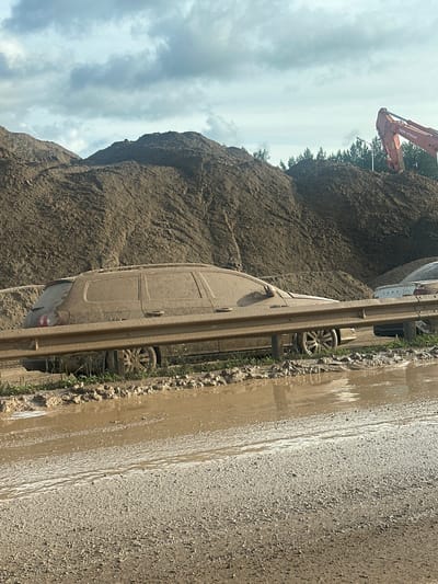 The image depicts a construction site in Khlystovo, Russia, under a cloudy sky, likely in the daytime. The foreground is dominated by a muddy road with a shallow pool of water reflecting the overcast sky. The ground is a mix of gravel and mud, with some sparse green grass peeking through along the edge of the road.

A light-colored van, heavily covered in mud, is parked alongside the road. The van appears to be a panel van with sliding doors and a rear hatch. Its wheels are also caked in mud. Next to the van is a metal guardrail, also partially obscured by mud and debris.

In the background, a massive pile of dark, wet earth rises steeply. Atop this pile, an orange excavator with the "TATA HITACHI" logo visible on its arm is at work. The excavator's arm is extended, suggesting it is in the process of moving or excavating the dirt. Behind the pile of earth, a line of trees is visible against the sky.

The overall scene suggests recent rain or a very wet construction environment, with vehicles and the surrounding landscape significantly affected by mud and dirt. There are no people visible in the image. The atmosphere is industrial and appears to be part of an ongoing construction or excavation project.