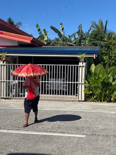 The image displays a bright, sunny daytime scene on a concrete road, consistent with a residential area in Burgos, Philippines. In the foreground, a person walks from left to right across the gray concrete pavement, casting a distinct, dark, oval-shaped shadow to their right. The individual's head and upper face are obscured by a large, open, red umbrella, patterned with white, yellow, and green floral or sunburst designs. They wear a horizontally striped shirt (white with red/pink stripes), dark blue shorts, and brown flip-flops. An orange fabric, appearing to be a scarf or face covering, is visible around their neck. A small, red, soft-material bag hangs from their left hand.

In the mid-ground, a white, vertical-barred metal gate is closed, forming part of a concrete perimeter fence. Behind this gate, a white vehicle, specifically a Mazda hatchback/small SUV, is parked within a garage or carport; its rear license plate "ZG 3B84" is partially visible. The structure covering the garage features a blue corrugated metal roof with white fascia. A small potted plant with green leaves rests atop a concrete pillar adjacent to the gate. To the right of the gate, dense green tropical foliage, including large-leafed plants, grows at ground level.

The background reveals parts of two residential structures. The one on the left has a red-orange corrugated roof and white eaves. The building housing the garage has a blue corrugated metal roof. Above and behind these structures, a dense canopy of tall, green banana trees with large leaves is prominent, interspersed with other tropical foliage and palm trees. The sky is clear and bright blue, indicating strong sunlight. A thin, dark utility wire stretches horizontally across the upper portion of the frame.