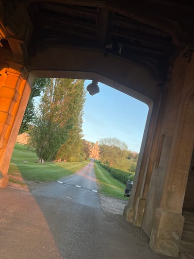 The image presents a view looking out from within a grand stone archway, showcasing a pathway leading to a distant stately building. The archway, carved from dark stone, frames the scene, with its intricate details and robust construction evident. The interior of the archway is largely in shadow, but its lower right side catches warm, golden sunlight, highlighting the texture and color of the stone.
Beyond the archway, a long, straight asphalt driveway or path stretches into the distance, flanked by expansive, well-maintained green lawns. A prominent, tall tree with dense green foliage stands to the left of the path in the midground. Further along the drive, rows of trees and hedges line the route.
At the vanishing point of the path, a large, ornate building, characteristic of a historic stately home or castle, is visible. It features multiple architectural elements, including numerous windows, suggesting a grand scale. The sky above is clear and bright blue, indicating fair weather. The lighting conditions, with warm, low-angle sunlight illuminating the scene and creating some shadows, suggest the time of day is either late afternoon or early morning.
A small, dark vehicle, possibly a utility cart or car, is partially visible on the right side of the path in the midground. A tiny dark shape, possibly a bird, can be seen in the sky. No people are present, and no specific activity is occurring, contributing to a serene and majestic atmosphere. No text is visible in the image. The architectural style and expansive grounds are consistent with a large estate found in areas like South Kesteven, United Kingdom.