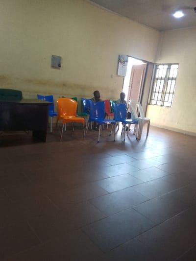The image depicts an indoor waiting area or common room in Nibo, Nigeria. Two males are seated in the mid-ground on blue plastic chairs, positioned with their backs partially towards the viewer. Both wear light-colored tops and dark trousers, with their gazes directed upwards and to the right.
The room features a dark brown tiled floor laid in a staggered rectangular pattern. The light cream-colored walls display a distinct horizontal band of discoloration across their lower third. A small, grey, square fixture is mounted on the left wall. An open, light-colored doorway is visible on the right wall, adjacent to which hangs a calendar. To the right of the doorway, a window with vertical and horizontal metal grilles allows natural light into the space.
A dark, rectangular desk or table is partially visible on the far left, with a dark green object resting upon it. Numerous unoccupied plastic chairs with metal legs are present in the mid-ground, exhibiting orange, green, red, blue, and white colors. The light-colored ceiling contains a single recessed light fixture. Illumination is provided by natural light entering through the window and doorway, supplemented by the artificial overhead light.