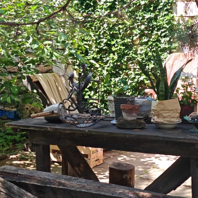 The image depicts a rustic outdoor setting, likely a garden or courtyard, within Останинское сельское поселение, Ukraine. In the foreground, a sturdy, dark-stained wooden table occupies the lower half of the frame. Its surface is adorned with various objects. From left to right, these include a silver-colored metal pot with a wooden handle, a dark, ornate metal candelabra or sculptural piece, and a black wire mesh bowl containing light-colored spherical objects. Further to the right are a partially obscured white container wrapped in a black mesh bag, a small terracotta pot with a plant, a pine cone, and a Sansevieria (snake plant) in a pot wrapped in crumpled brown paper, resting in a white saucer. A portion of a blue-green decorative dish with a rounded object is visible on the far right.
Under the table, wooden support beams are visible, along with two cut wooden stumps acting as possible seats or supports. On the ground beneath the table, a cardboard box with partial text (e.g., "AS", "ic") is present. The ground itself is composed of concrete or packed dirt, exhibiting dappled sunlight and shadows from overhead foliage. A dark wooden railing or bench partially crosses the very bottom of the image.
The background features dense green foliage. On the left, a stone wall is partially covered with broad-leafed plants, and a light-colored fabric draped over a wooden structure is visible. A blue plastic crate containing green plants is situated below this area. The central background is dominated by a vibrant green, densely grown ivy-covered wall. To the right of the ivy wall, a reddish-orange woven or mesh material is visible, possibly a screen or part of a structure, with white flowers in a dark pot in front of it. Overhead, tree branches with both broad leaves and needle-like foliage (possibly pine) cast shadows, contributing to the bright, sunlit outdoor ambiance. No people are present in the scene.