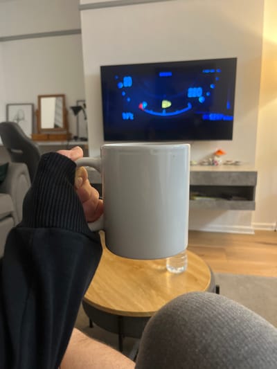 A person holds a large, white mug in front of a television screen displaying blue graphics, potentially a video game interface. A water bottle sits on a round, light-colored wooden table nearby. The individual is wearing a black sweater with ribbed cuffs. The setting appears to be a living room, with a gray couch, a decorative shelf, and wall art visible in the background.
