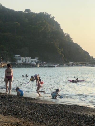 This image captures a late afternoon or early evening scene on a pebbled beach and bay in Sestri Levante, Italy. The prominent background features a large, steep, densely vegetated hill or promontory, likely overlooking the Baia del Silenzio or Baia delle Favole. At the base of the hill, across the bay, a multi-story white building complex with arched windows and terraces is visible, with a road and several parked vehicles in front of it. Numerous small boats are moored in the calm bay waters, demarcated by yellow buoys.
In the foreground, the pebbled beach transitions into the shallow water. A woman wearing a dark bikini stands in the ankle-deep water, viewed from behind. To her right, three children are actively playing at the water's edge: one child in a blue swimsuit kneels on the wet pebbles; another child, wearing a light-colored hat and swimsuit, runs through the shallow water, creating splashes, while holding a long, multi-colored water toy; and a third child in a light blue swimsuit crouches in the water with a blue bucket or scoop. Further out in the bay, several other individuals are present: two people are partially submerged in the water, and two others are on a pink inflatable raft, with another person nearby. The lighting suggests a low sun, casting soft light on the water and silhouetting the foreground figures against the brighter bay.