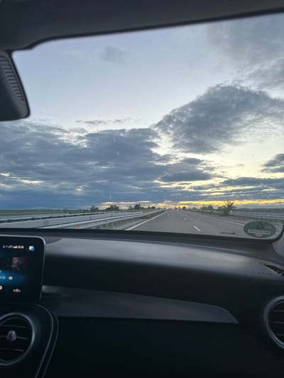 The image, taken from inside a car, shows a view of a highway in the city of Dobri Dol, Bulgaria. The road stretches into the distance under a sky filled with clouds. The car's dashboard and air vents are visible in the foreground. A small screen in the vehicle is also visible.