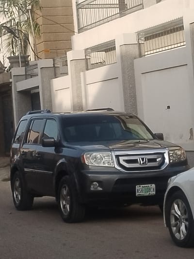 This image depicts a street scene in Unguwa Uku Kauyen Alu, Nigeria, under daylight conditions. The primary subject is a dark grey Honda Pilot SUV, parked on the left side of a paved road, facing towards the viewer's right. The vehicle displays a Nigerian license plate, "LAGOS NSR-108CN," mounted on its front bumper. It features roof rails, a visible sunroof, a chrome grille with the Honda emblem, clear headlights, and fog lights. To the right of the Honda Pilot, a portion of a white vehicle, showing its front wheel and bumper, is partially visible, indicating another parked car. In the background, a substantial building or compound wall extends horizontally, constructed of light-colored, possibly off-white, textured concrete. This wall is punctuated by several vertical pillars of the same material and features metal grates or fencing above sections. On the far left, a multi-story section of the building with a light brown textured facade and more metal railing is visible, along with palm tree foliage. No individuals or specific actions are discernible within the frame.