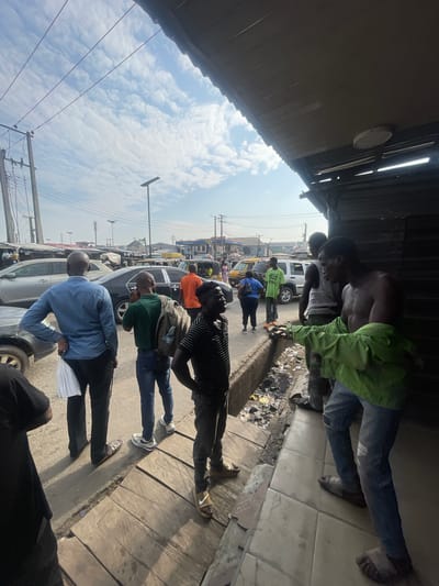 The image depicts a street scene in Akowonjo, Nigeria, likely a commercial area with a mix of parked vehicles, pedestrians, and market stalls. The sky is partly cloudy. Several individuals are visible. On the right side, two men are standing near a weathered structure, one of whom is shirtless and appears to be holding a green jacket. To the left, a man in a blue long-sleeved shirt is facing away from the camera, holding a white plastic bag. Another man in a green shirt with a backpack is also visible. Further in the background, various cars and a yellow bus are parked or in motion on the road. The ground consists of a combination of pavement, wooden planks, and a drainage ditch filled with debris. The overall impression is that of a busy, informal urban environment.