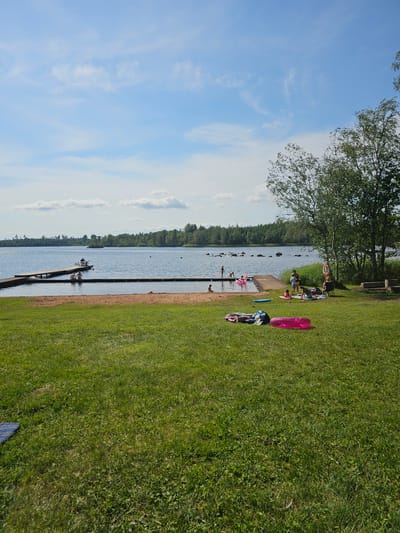 The image depicts a sunny day at a lakeside beach area, likely located in or near Smålandsstenar, Sweden, given the presence of typical Swedish summer scenery. In the foreground, a lush green lawn stretches out, dotted with a few inflatables, including a bright pink one, and a blue and white striped mat. Towards the middle ground, a sandy beach transitions into the calm, blue water of a lake. Several people are visible enjoying the water. A wooden pier extends into the lake, with a few individuals sitting on its edge. Further out on the lake, more people are swimming and playing. The background reveals a dense, green forest lining the far shore of the lake, with a few scattered rocks visible in the water near the shore. The sky above is a clear blue, with a few wispy white clouds, suggesting fair weather. On the right side of the image, near the edge of the lawn and closer to the trees, a group of people are gathered, some sitting on blankets, possibly picnicking or relaxing. A life preserver is visible hanging on a tree, hinting at a designated swimming area.
