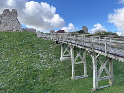 The image presents an outdoor landscape featuring ancient stone ruins on a grassy hill, accessed by a long, ascending wooden bridge. The bridge, constructed from weathered timber, has robust supports and railings, guiding the viewer's eye upwards towards the hilltop. The hill itself is covered in vibrant green grass, interspersed with patches of lighter earth and sparse vegetation.
Dominating the crest of the hill are extensive ruins of a castle or fortification, characterized by rough, grey stone walls, likely flint. A prominent ruin, possibly a tower, is visible on the left side of the hill, with other fragmented walls and a more distant circular tower further along the ridge.
The sky above is a bright blue, adorned with numerous large, fluffy white cumulus clouds, indicating clear and pleasant weather conditions, likely mid-morning or afternoon. No people are visible, and no specific activity is occurring, contributing to a tranquil and historical atmosphere. Given the provided location context of King's Lynn and West Norfolk, United Kingdom, these ruins are consistent with those of Castle Rising, known for its distinctive flint architecture. There is no visible text in the image.
