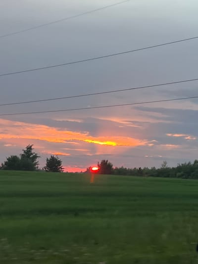 The image depicts a sunset over a rural landscape, likely in the vicinity of Поварёнки, Russia. A vast expanse of green field stretches across the foreground, with a line of trees forming a distant horizon. Above the trees, the sky is a canvas of dramatic colors. A band of vibrant orange and yellow clouds is illuminated by the setting sun, which appears as a bright red orb peeking through a break in the clouds. Faintly visible in the upper portion of the image are power lines, suggesting a proximity to infrastructure. The overall impression is one of natural beauty and tranquility, with the dramatic sunset providing a focal point against the serene rural backdrop. There are no people or animals visible in the image.