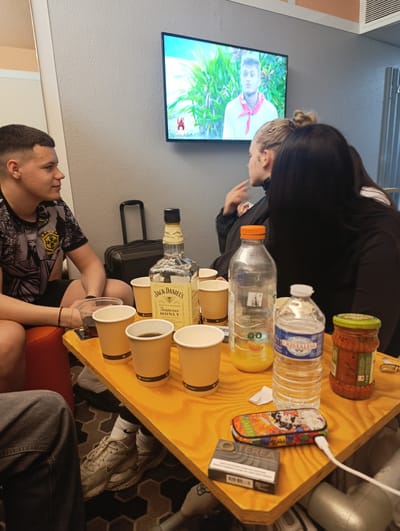 The photograph depicts a casual indoor scene, likely in a residence or hotel room in Bobigny, France. Three individuals are partially visible, focused on a television mounted on the wall. A young man sits on an orange chair, glancing towards the television. A young woman with blonde hair in a bun appears to be speaking or gesturing. Another individual is partially visible, her back turned, and wearing dark clothing. A luggage bag is nearby, and an air conditioning unit can be seen in the upper right corner.
A small yellow table is positioned in the center of the room. On the table are several cups filled with liquid, a bottle of Jack Daniels Honey whiskey, bottles of orange juice and water, a jar of sauce, a cigarette box, and a colorful charging case with a white cable connected to it.