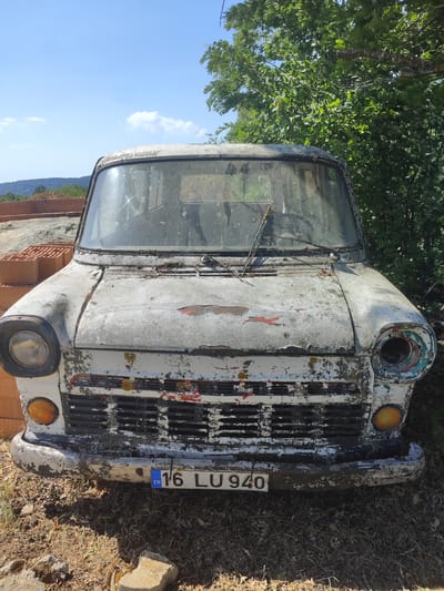 The image shows the front of a heavily weathered and disused white van, likely parked outdoors in a rural or semi-rural setting. The van is very dirty, with peeling paint revealing rust and other colors underneath. The front grille is intact but also shows signs of decay. One of the headlights is present, while the other is missing its lens, exposing the internal components and a colorful, decorative ring around the opening. Orange turn signals are visible below the headlights. The windshield is dusty and smudged, obscuring the interior. Two windshield wipers are present, one resting on the glass and the other slightly lifted.

In the foreground, dry leaves, dirt, and some rocks cover the ground. Behind the van, a stack of orange bricks is visible, suggesting construction or renovation activity nearby. Further back, lush green trees and foliage dominate the background, with a clear blue sky overhead dotted with a few white clouds. A distant hillside is visible on the left.

The overall impression is one of abandonment and the passage of time. The bright sunlight and clear sky suggest it is daytime, likely a warm and dry day. The image captures a sense of neglect and decay in contrast with the surrounding natural beauty.

Visible text on a license plate on the front of the van reads "TR 16 LU 940". This indicates the vehicle is registered in Turkey, specifically the province of Bursa (represented by the number 16). The context of Osmangazi, Turkey, aligns with this observation.