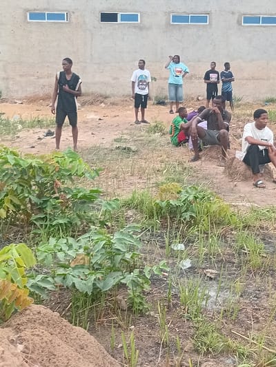 A group of people are gathered outside near a building with windows. Some are standing, while others are sitting or crouching on the ground. A few individuals are wearing t-shirts, and one is dressed in white. Vegetation is visible in the foreground, including plants and grassy areas. The setting appears to be a casual outdoor environment, possibly near a residential or community building.