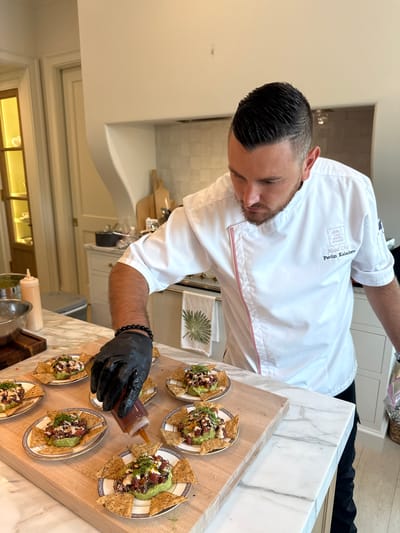 A male chef, identified as Pavlin Kalachev, Head Chef at The Rose House, is shown plating small appetizers in a kitchen located in East Naples, United States of America. He wears a white chef's coat with pink trim and black gloves, meticulously drizzling sauce from a squeeze bottle onto a dish. The dish consists of what appears to be a scoop of avocado topped with diced tuna or salmon, garnished with herbs and drizzled with sauce, served on a round plate with a decorative blue and white border, surrounded by tortilla chips. Several other identical appetizers are already plated on a wooden cutting board, suggesting a catering or restaurant preparation setting. The kitchen environment features marble countertops, white cabinetry, and stainless steel appliances visible in the background.