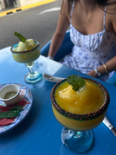 The image shows a blue table with two frozen cocktails and a small bowl of green salsa. The table is set on an outdoor patio, with a blurred background of a street. On the table, a small plate with red and green chips rests next to the green salsa. The frozen cocktail on the right has a yellow base, rimmed with red chili powder. A single green mint leaf sits atop the drink. The frozen cocktail on the left has a green base and a brown rim. Both cocktails are served in clear glass cups with frosted blue rims. A woman wearing a white dress and sitting in a blue chair is visible in the background, with her right hand resting on the table. The scene is located in Pozos, Costa Rica, at the coordinates 9.951 latitude and -84.194 longitude.