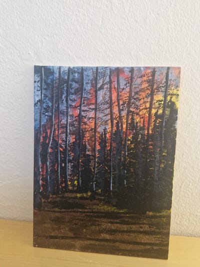 The image displays an acrylic painting on canvas depicting a forest scene at sunset. The sky is ablaze with hues of orange, yellow, and red, visible through the vertical trunks of dark trees. The foreground features a textured ground, likely meant to represent earth or grass, with patches of yellow and green suggesting scattered vegetation. The painting is positioned against a textured white wall, with a wooden surface visible at the bottom of the frame. There is no indication of the city of Новоангарск, Russia, within the image itself. The painting is the sole subject of the photograph.
