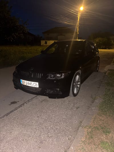 A black BMW wagon is parked on a paved road at night. The car's license plate reads "BP 2445 CB". A street light illuminates the scene, casting long shadows and highlighting the car's features. The background shows houses and trees, suggesting a residential area. The overall environment is dark, with a clear night sky.