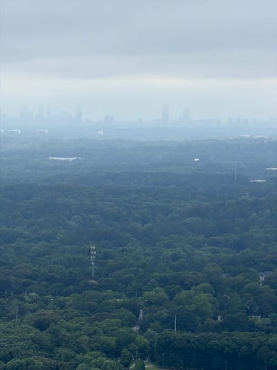 An aerial view showcases Stone Mountain, United States. The scene features dense green forestation to the right, blurring into a light hazy mist to the left, where indistinct city skylines are faintly visible. Power lines run parallel to the dense forest. The image appears to be taken from a high vantage point, possibly an elevated building or drone.