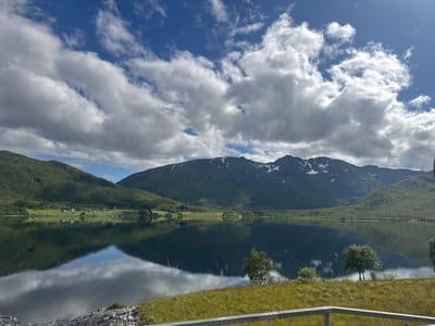The image captures a serene landscape in Gullesfjord, Norway, dominated by a calm lake reflecting the sky and surrounding mountains. The sky above is a vibrant blue, dotted with fluffy white clouds. Majestic mountains, partially covered in snow, rise in the background, their slopes carpeted with lush green vegetation. The foreground features a grassy embankment with a few scattered trees and a rocky shoreline. In the distance, several houses are nestled amongst the green fields, suggesting a small settlement. The overall impression is one of tranquility and natural beauty.