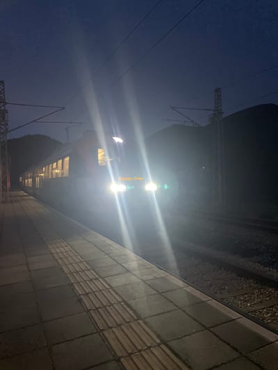 The scene depicts a train at a station in Vlado Trichkov, Bulgaria, during nighttime or deep dusk. A passenger train, likely an electric multiple unit, occupies the center-right of the frame, brightly illuminated by its two powerful front headlights, which generate significant lens flare. A destination sign between the headlights displays "Банкя - София" (Bankya - Sofia). The train's interior lights are visible through its windows along the left side.
To the left, a light-colored paved station platform extends into the foreground, featuring a strip of tactile paving with raised ridges running parallel to the tracks. Two sets of railway tracks are visible to the right of the platform, supported by ballast. Overhead catenary wires and their support structures, including a prominent lattice mast on the right, are present above the tracks. The background reveals dark, indistinct mountain silhouettes against a dark blue sky, indicating the surrounding mountainous terrain. No individuals are visible.