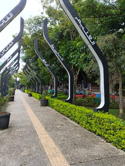 A long walkway with a tactile paving strip stretches into the distance, flanked by identical curved, black and white structures. Each structure bears Arabic script. Manicured green hedges line the path, with occasional planters along the side. Trees provide a canopy overhead, filtering the light. In the background, glimpses of buildings and activity can be seen.