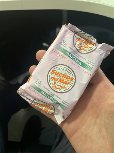 A hand is holding a sealed, light purple, single-serve Alfajores de Maicena cookie. The packaging includes the brand name “Sueños del Mar”, the weight “50g”, and the description "Alfajor relleno con dulce de leche y coco rallado MAICENA Elaborados en la costa Industria Argentina". It cannot be determined from this photo if this product is specifically located in the city of Piñeyro, Argentina.