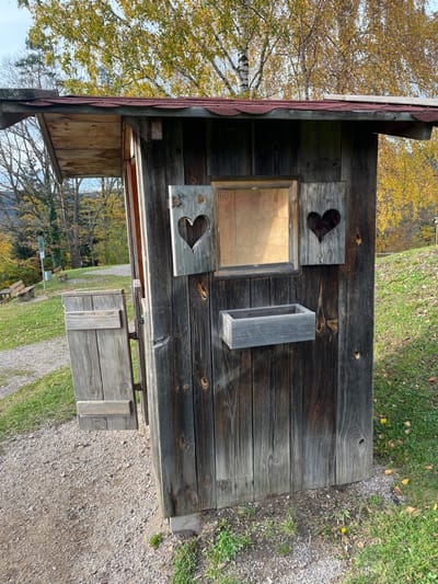 The image displays a weathered wooden hut or kiosk situated outdoors in a scenic area of Oppenau, Germany. The structure features a dark-stained vertical wood plank exterior and a red shingle roof with an overhang. The front facade contains a central, square, glass-paned window that appears to display a faded document. This window is flanked by two light-grey wooden shutters, each with a heart-shaped cutout; the left shutter is slightly ajar. Below the window, a rectangular wooden planter box is affixed to the wall. On the left side of the hut, a smaller, light-grey wooden door or panel is ajar, opening outward. The hut rests on visible concrete or stone foundation blocks.

To the left of the hut, a gravel path extends into the background, where a green signpost and a wooden bench are visible. Green grass covers the ground to the right of the hut and borders the path, with scattered yellow leaves present. In the background, autumnal deciduous trees with yellow and gold foliage are prominent, including a birch tree with white bark behind the hut's roof. Distant hills covered with both deciduous and evergreen trees are discernible under a clear sky. No individuals or active subjects are present in the scene.