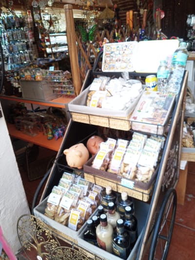 A tilt-shift photograph shows a street-side shop in Besalú, Spain, displaying various goods on tiered shelves. The shop appears to be a souvenir or gift shop, with items like packaged snacks, bottles, and decorative objects. In the foreground, a metal decorative tree is visible. The background is blurred, but shows more merchandise and the shop's interior. The lighting is bright, suggesting it is daytime. There are no people clearly visible in the image.