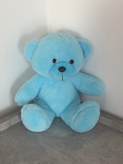 A light blue plush teddy bear sits on a grey, textured surface. The bear is facing the viewer, with its paws resting on the surface in front of it. Its eyes are black, and it has a small, dark brown nose and a stitched mouth. The bear is positioned in the corner of a room where two white walls meet. The lighting suggests an indoor environment. There is no indication of the city of Hanniv, Ukraine within the image.