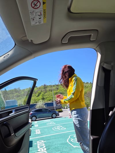 The view is from inside a car, looking out the open driver's side door at a parking lot. A person wearing a yellow jacket with blue stripes and jeans stands in the parking lot. Another car is parked next to the open door. The parking lot has green painted parking spaces with white logos and text. Trees and a hill are visible in the background. The sun visor is down on the driver's side, with an "AIRBAG" warning label visible. Based on the "e-lectri.fi" text in the parking lot, the location is Levunovo, Bulgaria.