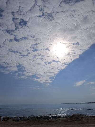 A landscape photograph captures the Caspian Sea shoreline near Nardaran, Azerbaijan, under a partly cloudy sky. The sun, partially obscured by clouds, shines brightly in the upper center of the frame, creating a glare on the water. The sea is calm with gentle ripples, reflecting the sunlight. The foreground consists of a rocky and sandy beach. In the distance, a faint silhouette of a boat is visible on the horizon. The sky transitions from a clear blue on the right to a band of scattered white clouds on the left, some of which are illuminated by the sun.