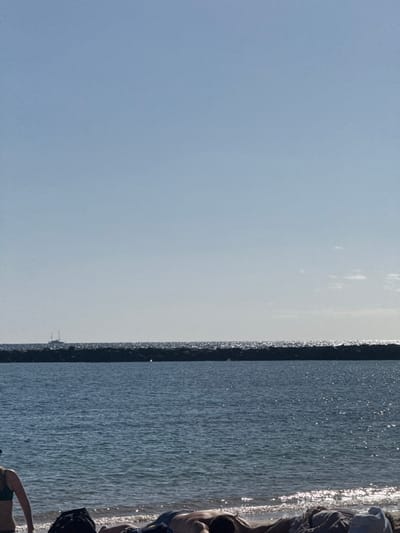 The image captures a bright, sunny coastal scene in Los Cristianos, Spain, with a clear blue sky dominating the upper left portion of the frame. The horizon line, marked by a dark, thin breakwater or jetty, stretches horizontally across the center of the image, separating the expansive sky from the sparkling, calm ocean water below.
The water surface is covered in numerous small glints of sunlight, indicating a bright overhead sun. On the far right side of the image, a sandy or pebbled shore is visible, where several individuals are partially seen. One person's torso and arm, seemingly clad in dark swimwear, are prominent near the water's edge, suggesting they are relaxing or sunbathing. Other indistinct figures are also visible, either sitting or lying down on the beach, enjoying the sunny weather. A small, distant structure, possibly a boat or part of a pier, can be vaguely discerned near the horizon line on the left side of the breakwater. The overall impression is one of a peaceful and leisurely day at the beach. No visible text is present in the image.