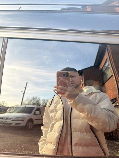 The image displays a male individual's reflection taking a selfie in a vehicle window, situated in Bretton, United Kingdom. The man, seen from the chest upwards, wears a light-colored, possibly cream, puffer jacket with black trim along the zipper. He holds a smartphone with a light orange-pink case, featuring a circular MagSafe ring, in his right hand, partially obscuring his face. His hair is short and dark with faded sides, complemented by short facial hair.
The reflected background reveals an outdoor environment under a clear blue sky with scattered wispy clouds. To the left of the individual, a white Vauxhall Corsa van is parked on an unpaved surface; its partial UK registration plate, "CA58 BXE," is visible. A utility pole with several cross-arms stands further to the left, alongside bare trees in the distance. To the right, a multi-story brick building with dark window frames and a dark roofline is partially reflected. The scene is illuminated by strong, low-angle sunlight, suggesting either early morning or late afternoon.