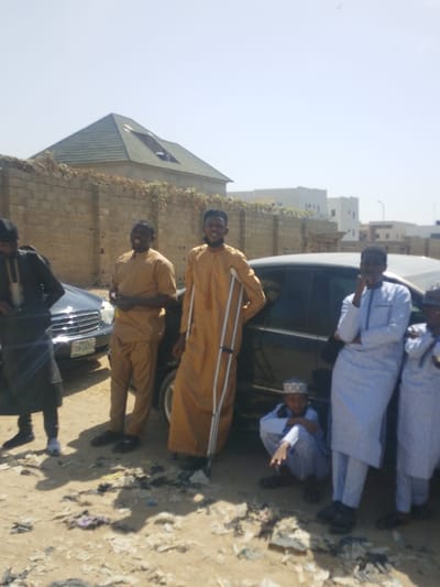 In Darmanawa, Nigeria, an outdoor scene depicts seven males standing and squatting on unpaved, dusty ground under a clear, bright sky. Two black sedans are positioned behind them.
From left to right:
1. A male in dark traditional attire stands near the front of a black Mercedes-Benz sedan, partially obscured. A Nigerian license plate is partially visible on the vehicle.
2. A male in a tan traditional tunic and trousers stands smiling, looking towards the right.
3. A male in a tan kaftan, wearing a dark cap and sporting a beard, stands smiling towards the camera, supporting himself with silver aluminum crutches under his left arm. He is positioned beside a second black sedan.
4. A young male, dressed in a light blue traditional tunic, trousers, and cap, squats on the ground in the foreground, looking towards the camera.
5. A male in a light blue traditional tunic and trousers stands leaning against the second black sedan, with his right hand raised near his face.
6. A second male, partially visible, wearing similar light blue attire and a cap, stands next to the fifth male, also leaning against the car.
The immediate ground surface is light-colored dirt with scattered pieces of debris and trash. In the background, a light-brown brick wall is visible, topped with green foliage. Behind the wall to the left, a building with a green pyramidal roof is observed. Further in the background to the right, several modern-style white buildings are present.