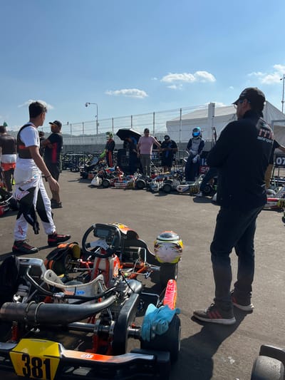 The image depicts a gathering of people and go-karts in an outdoor setting, likely a racing event. The location appears to be in Amozoc de Mota, Mexico, based on the presence of a go-kart with the number "381" and "www.elischart.com" on it, which could refer to a karting organization or team based in that region.
Several individuals are visible, including a man in a black shirt and cap, who seems to be attending to one of the karts. Another man, wearing white racing attire, is standing near a kart with the number "381". Other people are scattered throughout the scene, some of whom are observing the karts.
The primary subjects are the go-karts, which are lined up and appear ready for a race or practice session. The karts have various designs and colors, some bearing numbers and logos. A helmet is placed on one of the karts, and gloves are also visible.
The environment suggests a sunny day at a racing venue, with a paved surface and a fence in the background. A partially visible tent or structure is present in the upper part of the image.