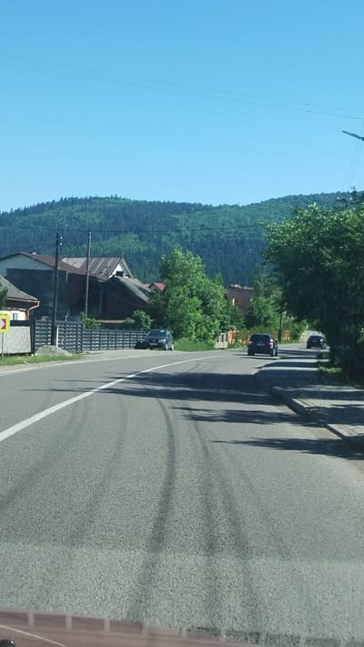 This is a landscape-oriented, slightly tilted, first-person view from a vehicle traveling along a road in a rural, mountainous region. The road is paved and has a white line marking the lane. The scene is characterized by a dense, dark green forest covering a steep mountainside that dominates the left side of the frame. The sky is a clear, bright blue.
Several cars are visible on the road ahead, all appearing to be dark in color and moving in the same direction. There are some houses and buildings visible at the base of the mountain, interspersed with trees. The architecture of these buildings appears to be typical of a European rural setting, with dark roofs and wooden elements. One building has a dark, tiled roof and a gray facade. Another has a lighter colored roof and a stone-like lower level. A yellow diamond-shaped sign with a black border and possibly a black symbol is visible near the bottom left of the frame.
On the right side of the road, there are shadows cast by trees and possibly a utility pole, creating a striped pattern across the asphalt. The overall impression is of a sunny day, likely in the late morning or early afternoon, given the brightness and the direction of the shadows. The environment suggests a location in a natural, possibly temperate climate, with the visible greenery indicating a time of year like late spring or summer. The activity depicted is simply traveling by car through a scenic, wooded area with scattered human habitation. There is no clear indication of any specific event or emotional tone.