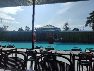A view from under a covered patio looking out onto a swimming pool in Gambiran, Indonesia (latitude: -8.434, longitude: 114.18). There is a blue swimming pool with a life preserver on the left and a "no diving" sign on the right. The pool is surrounded by a green hedge. There are several black wicker chairs and tables set up in front of the pool, the chairs are empty, and the tables have a few empty plates and napkins. The sky is clear and bright blue with a few puffy white clouds. There are several trees to the left and right, and a low building with a glass roof in the background. The patio is partially shaded and the light is bright. There is no one visible in the image.