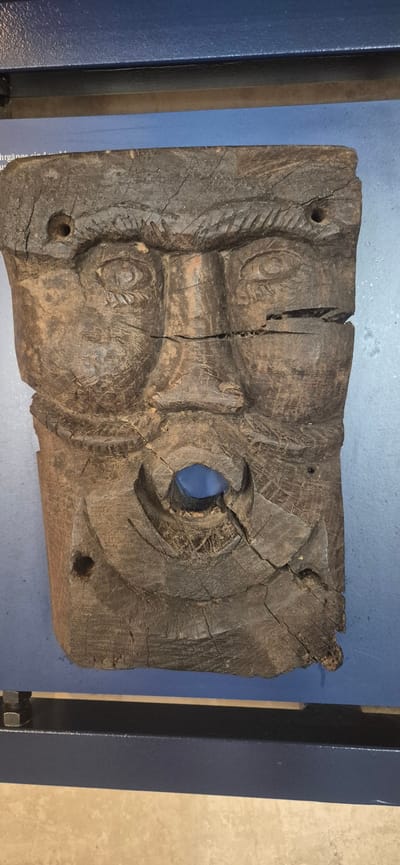 The image shows a close-up, vertical shot of a dark brown, ancient wooden mask. The mask is carved with the features of a face, including two prominent eyes with carved pupils and eyelashes, a nose, and a mouth with a round opening in the center. The wood is weathered and cracked, showing its age. There are two circular holes on the left side of the mask, one near the top and one near the bottom. The mask is positioned against a blue background, possibly a display wall in a museum. Below the mask, a dark metal frame with bolts is visible. The overall impression is of an archaeological artifact displayed for viewing.