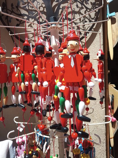 This is a close-up, eye-level, outdoor photograph of a display of wooden Pinocchio puppets hanging from a white metal rack. The puppets are predominantly red and feature wooden bodies, limbs, and heads with painted facial features and red conical hats. Some puppets have black hair and white shirts with black ties. Their limbs are jointed, allowing for movement. The puppets are strung together with red thread and are displayed in multiple rows. In the background, a textured wall with a decorative wrought-iron shadow of a tree is visible. To the right of the puppets, a wooden cutout of what appears to be a sword or shield is leaning against the wall. Further down, smaller wooden toys, including what look like colorful ladybugs and pigs, are also displayed. The lighting suggests it is daytime, with strong sunlight casting shadows. The scene appears to be a street vendor stall, likely in a tourist area of Besalú, Spain, given the typical souvenir offerings found in historic Spanish towns. There are no people in the frame.