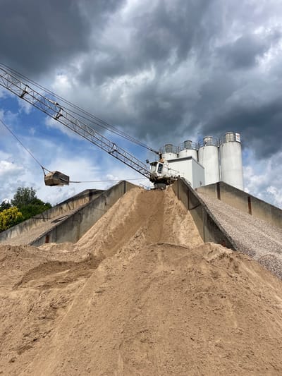 This is a medium-shot, exterior, ground-level, eye-level, vertical photograph of a construction or industrial site on a cloudy day. In the foreground, large piles of light brown sand dominate the scene. Behind the sand, a concrete wall slopes upwards, topped with a pile of gravel. Further back, a white industrial building with several tall, cylindrical silos is visible. A large, white crane with a long boom extends from the left side of the image, its bucket suspended in the air over the concrete wall. The sky is filled with dark, dramatic storm clouds, with patches of blue sky visible between them. The overall impression is one of industrial activity under a brewing storm. The location is an industrial area in Bielefeld, Germany, likely a gravel or cement plant. There are no people visible in the image.