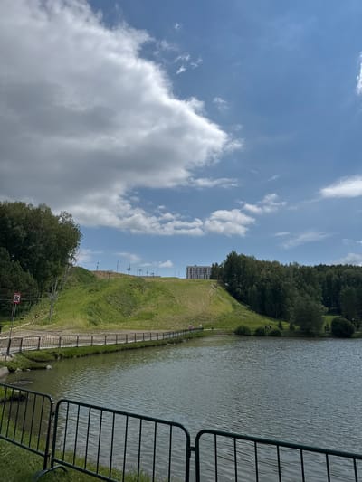 An outdoor scene depicts a large body of water in the foreground, bordered by a black metal fence. Beyond the water, a grassy hill rises, with a paved walkway running along its base, also separated by a black metal fence. A few individuals are visible walking on the walkway and the grassy slope. The hill is dotted with ski lift towers and is topped with trees and a multi-story building. The sky above is blue with scattered white clouds. The image is taken in Koltsovo, Russia.