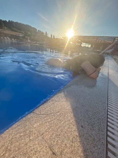 A person is resting at the edge of a swimming pool. The person is partially submerged in the bright blue water, with their head resting on their arms which are on the pool's edge. They are wearing dark clothing and a bracelet on their left wrist. The sun is shining brightly in the background, creating lens flare and casting long shadows. The pool area appears to be a resort or hotel, with lounge chairs and umbrellas visible in the distance, situated near a wooded hillside. The poolside is made of textured stone, and a slatted drain runs along the edge. The image evokes a sense of relaxation on a sunny day. The location is not specified in the image itself.
