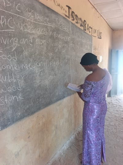 An adult female stands in profile within a classroom located in Mada, Nigeria. She wears a long, patterned purple dress with a black head covering and holds an open notebook, pointing to its pages with her right index finger. Her attention is directed downwards towards the book. To her left, a large dark grey blackboard occupies the upper portion of a light brown, textured wall. The blackboard displays white chalk writing, including "BASIC SCIENCE," the date "12/03/2026," and the topic "uses and Importance of Living and non-living things," followed by a list of items such as "Food," "Some medicinal plant," "Rubber," "Wood," and "Stone." Additional notes regarding "Living thing provide food" and "aloe-vera" are visible on the right side of the blackboard. The light brown wall above the blackboard bears black painted text, including "Jss2B ACTION CLASS." The floor to the right of the woman appears to be bare earth or sand. A bright light source, likely an open doorway or window, is visible on the far right.