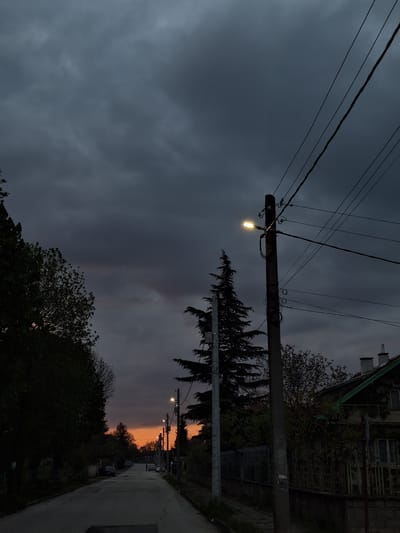 The image depicts a quiet street scene at dusk in Dolni Chiflik, Bulgaria. The sky is predominantly dark grey, filled with heavy, overcast clouds that cover most of the upper frame. However, a distinct strip of warm orange and red light is visible on the horizon towards the right, indicating the last moments of sunset.
Several utility poles are prominent, featuring numerous power lines that crisscross the frame. One streetlight, centrally located on a pole, is illuminated and glows with a warm yellow light, while other streetlights can be seen further down the road, also lit. The environment is rich with trees, including a tall, dark coniferous tree with distinctive foliage, and various deciduous trees, their silhouettes stark against the fading light.
A narrow, paved road or path recedes into the distance on the right side of the image, bordered by what appears to be houses and fences, largely obscured by shadow. One building with a gabled roof and windows is partially visible in the upper right corner, hinting at a dwelling. There are no people, vehicles, or specific activities visible in the frame. The scene conveys a tranquil and atmospheric mood, typical of twilight hours. No visible text is present in the image.