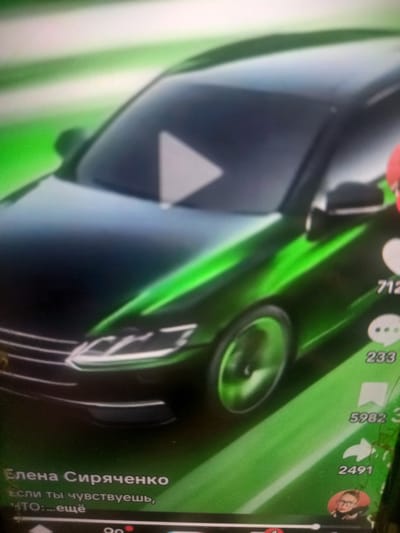The image is a blurry screenshot of a TikTok video featuring a car with a green metallic paint job. The video appears to have some kind of overlay, with a play button icon in the center of the car's windshield and social media interaction icons with numbers (712, 233, 5982, 2491) on the right side. Text is also visible at the bottom left corner: "Елена Сиряченко" and "Если ты чувствуешь," followed by what appears to be censored text "...ещё". The car is the primary subject, with its sleek design and vibrant green color dominating the frame. The background is blurred, with streaks of green and white suggesting movement or a stylized effect. There are no visible elements that would suggest a specific location in Maloye Lugovoye, Russia, or any other city. The image is of low resolution and suffers from significant motion blur, making precise identification of details challenging.