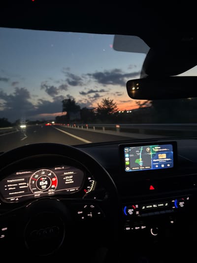 The image shows the interior of a car at dusk, with the dashboard and steering wheel in the foreground. A digital display shows the car is traveling at 84 km/h. The car is on a highway with other cars visible in the distance. The sky is a gradient of blue and orange, indicating sunset. The car is equipped with a navigation system showing a map and route. The climate control system is visible, set to 20 degrees Celsius. The scene suggests the car is in motion, likely on a road trip or commute during the evening. There are no people visible in the car. The location is specified as Ignatievo, Bulgaria, though this is not visually verifiable from the image itself.