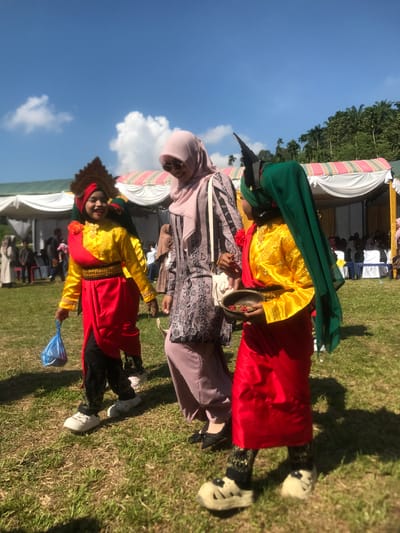 In Pantonrayeuk Satu, Indonesia, three individuals are captured outdoors on a sunny day. The main subjects are two young girls in traditional attire, likely participating in a cultural event, walking across a grassy field. The girl on the left wears a yellow top with red accents, a red skirt, and a decorative headdress. She carries a blue plastic bag. The girl on the right is dressed in a yellow top with a red wrap and skirt, and a green headscarf. She holds a small bowl containing reddish seeds. Between them, an adult woman, veiled in a light pink hijab and wearing a patterned dress, walks alongside them, looking down and smiling. In the background, white tents are set up, suggesting an outdoor gathering or festival. Palm trees are visible in the distance, bordering the scene. The overall atmosphere appears festive and communal.