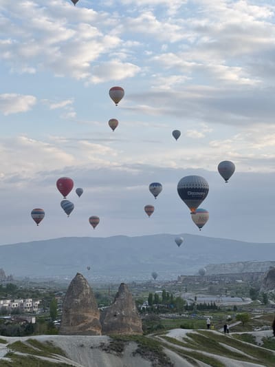 The image shows a sky filled with hot air balloons in Göreme, Turkey. The balloons are of various sizes and colors, some with company logos. In the background, there is a view of the landscape, featuring unique rock formations and buildings of the city, situated in a valley under a hazy sky. The horizon is indistinct due to the mist or haze.