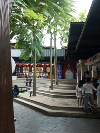 This image depicts an outdoor commercial plaza located in Tân Kỳ Commune, Vietnam. The scene is dominated by a multi-tiered concrete step system forming a terraced seating or circulation area. Several tall, slender trees with green foliage are centrally planted within the concrete structure. In the background, a building with a corrugated metal facade spans horizontally, featuring commercial establishments. One visible shopfront, possibly a gelato vendor, displays signs in English such as "ALL YOU GELATO" and "COLD, CREAMY, DREA...", characterized by illuminated square frames and a whimsical decor with yellow chairs. Further right in the background, a large white, textured spherical display is set against a red curtain. On the far right, a structure with a dark, sloped roof overhangs a retail display area showing multiple shelves with various items.
Multiple individuals are present. In the lower-left foreground, one person is seated on a curved concrete ledge, wearing a dark hooded jacket, blue jeans, and red footwear. In the midground-left, two adults are seated on a higher concrete ledge. On the right-midground, a group of approximately five individuals, including at least three children and two adults, are standing and congregating on the concrete steps. One child wears a striped long-sleeved shirt, another a light-colored dress, and a third a light blue patterned top. An adult in a dark top is visible amongst this group. The ground level is paved with interlocking gray paver stones. The lighting is diffused, suggesting an overcast day or indirect light conditions.